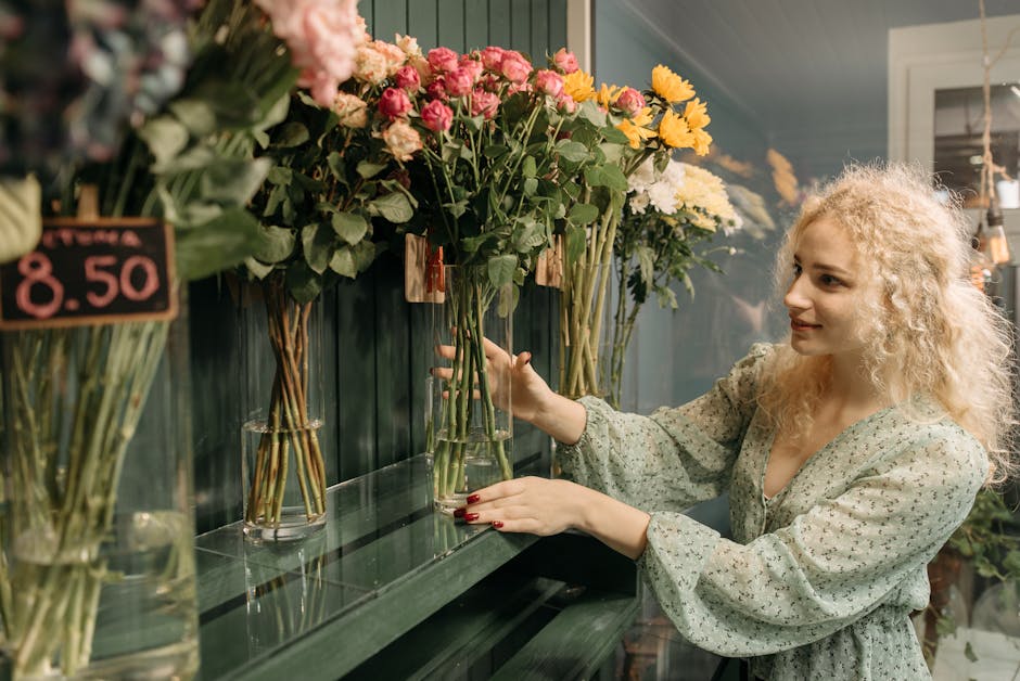 A woman with curly blonde hair selecting a flower bouquet from a display of fresh flowers inside a florist shop. She is wearing a light, patterned blouse and has neatly manicured red nails. The bouque