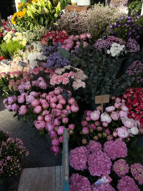 A vibrant display of fresh flowers at a florist shop, featuring pink peonies, purple chrysanthemums, and white lilies arranged in bunches and bouquets. The flowers are arranged in buckets and baskets,