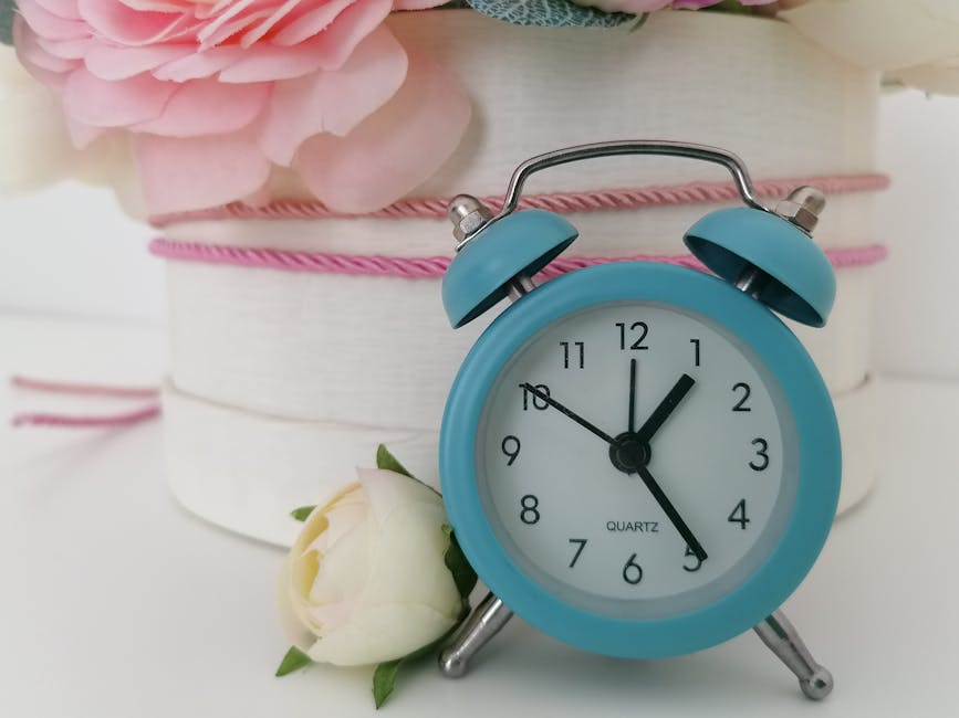 A light blue vintage-style alarm clock displaying the time as 12:05, with two bells on top and silver legs, positioned beside a single white ranunculus flower with green leaves at its base. The clock