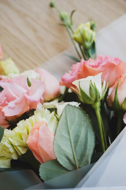 A close-up of a floral arrangement featuring pale pink and cream lisianthus flowers with ruffled petals, complemented by broad green leaves and small unopened buds. The bouquet is wrapped in white tis