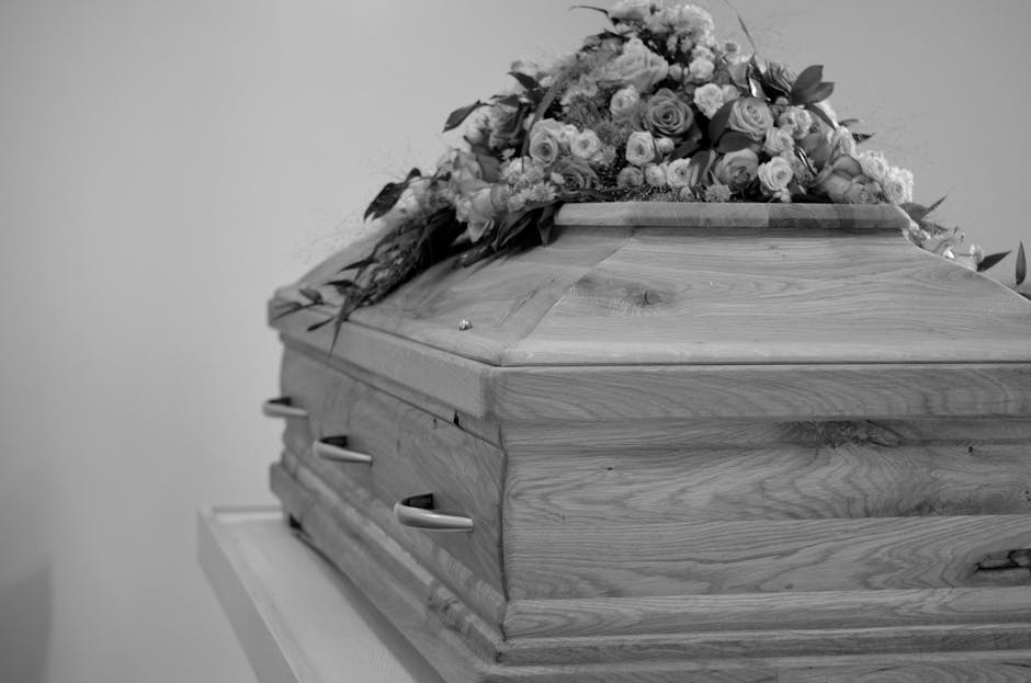 A wooden casket resting on a white stand, topped with a floral arrangement comprised of roses, lilies, and assorted blooms in soft pastel shades, wrapped in transparent cellophane and tied with a ribb