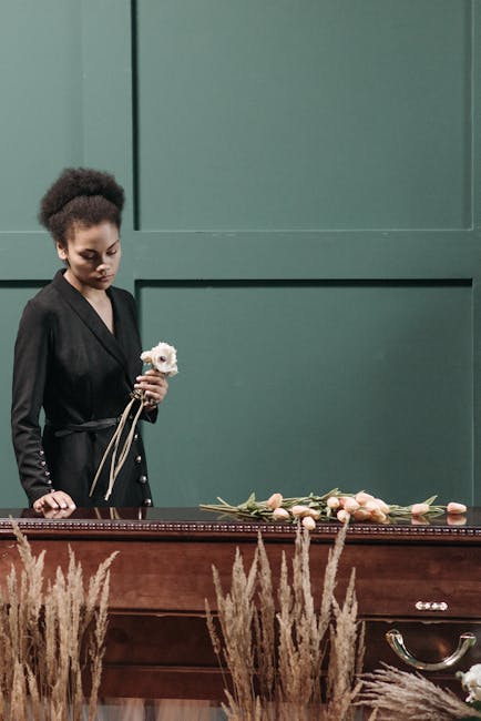 A young woman with dark curly hair styled in a natural updo stands beside a wooden funeral casket decorated with a floral arrangement of soft pink roses and greenery. She is dressed in a black blazer
