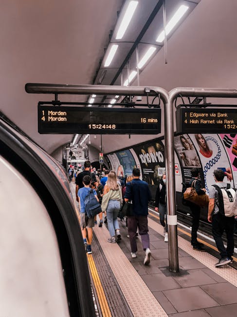 A vibrant and diverse group of commuters standing on a busy underground station platform, with some walking and others waiting near the edge. Overhead, illuminated electronic departure boards display