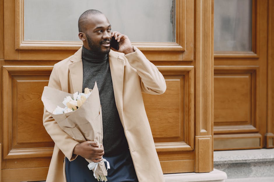 A man with dark skin and a neatly trimmed beard stands outdoors against a wooden-paneled building, holding a bouquet of fresh white flowers wrapped in brown paper. He is wearing a beige overcoat over